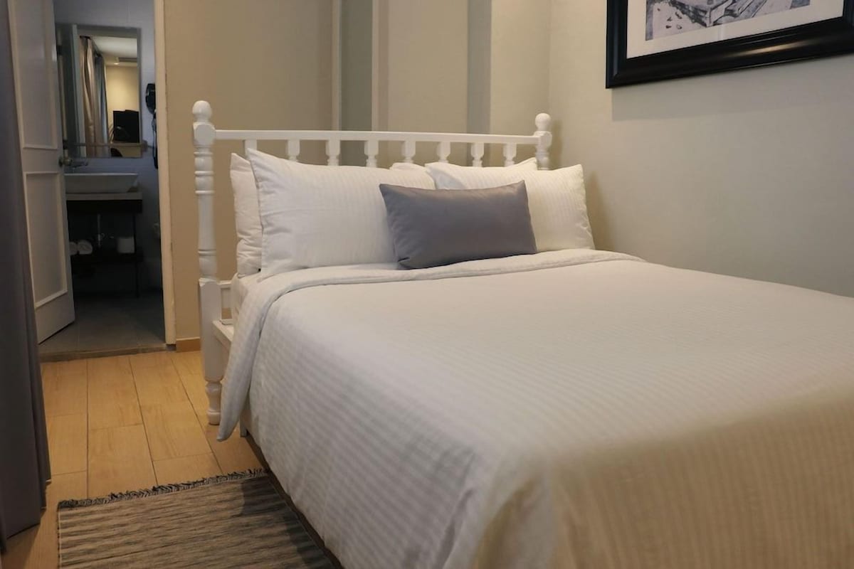 Una cama bien hecha con ropa blanca y una sola almohada gris en una habitación con cabecera blanca.