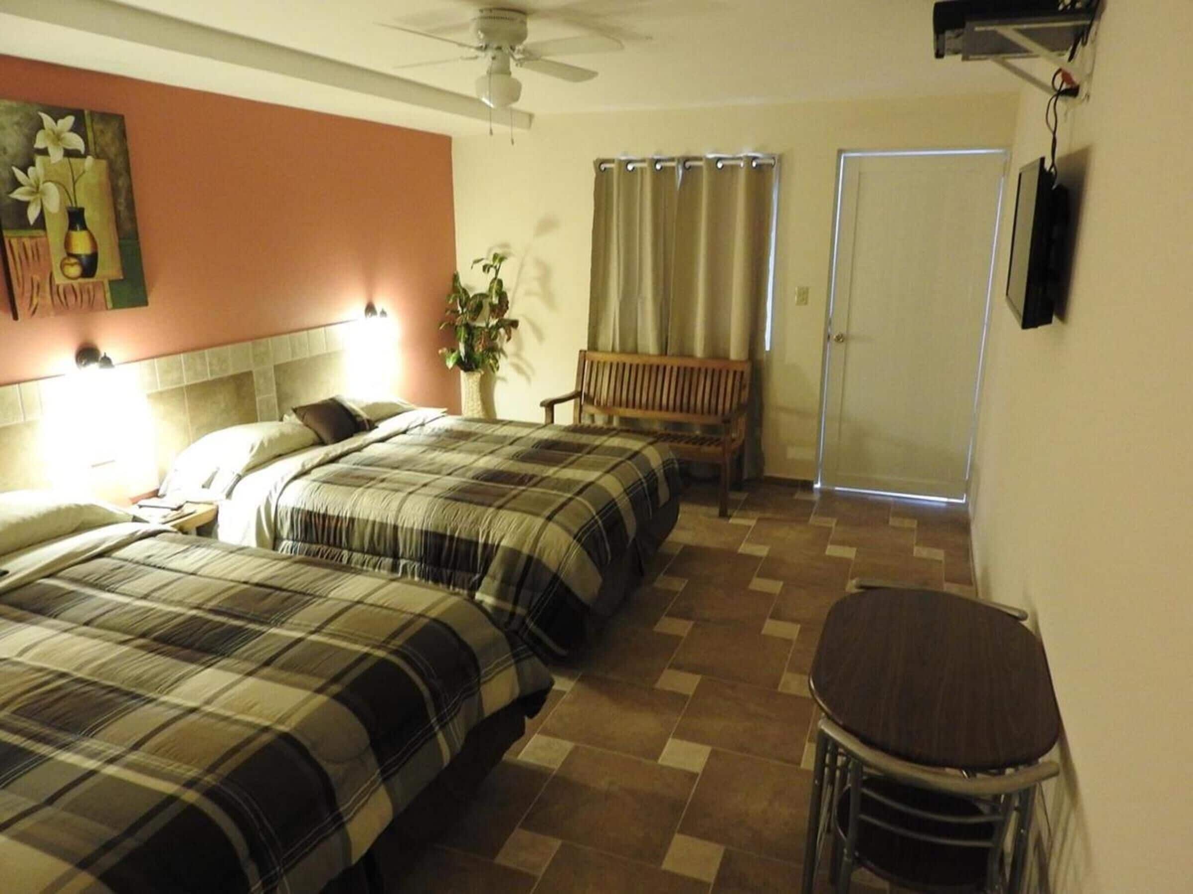 A hotel room with two beds, a bench, a table, a ceiling fan, and a door.