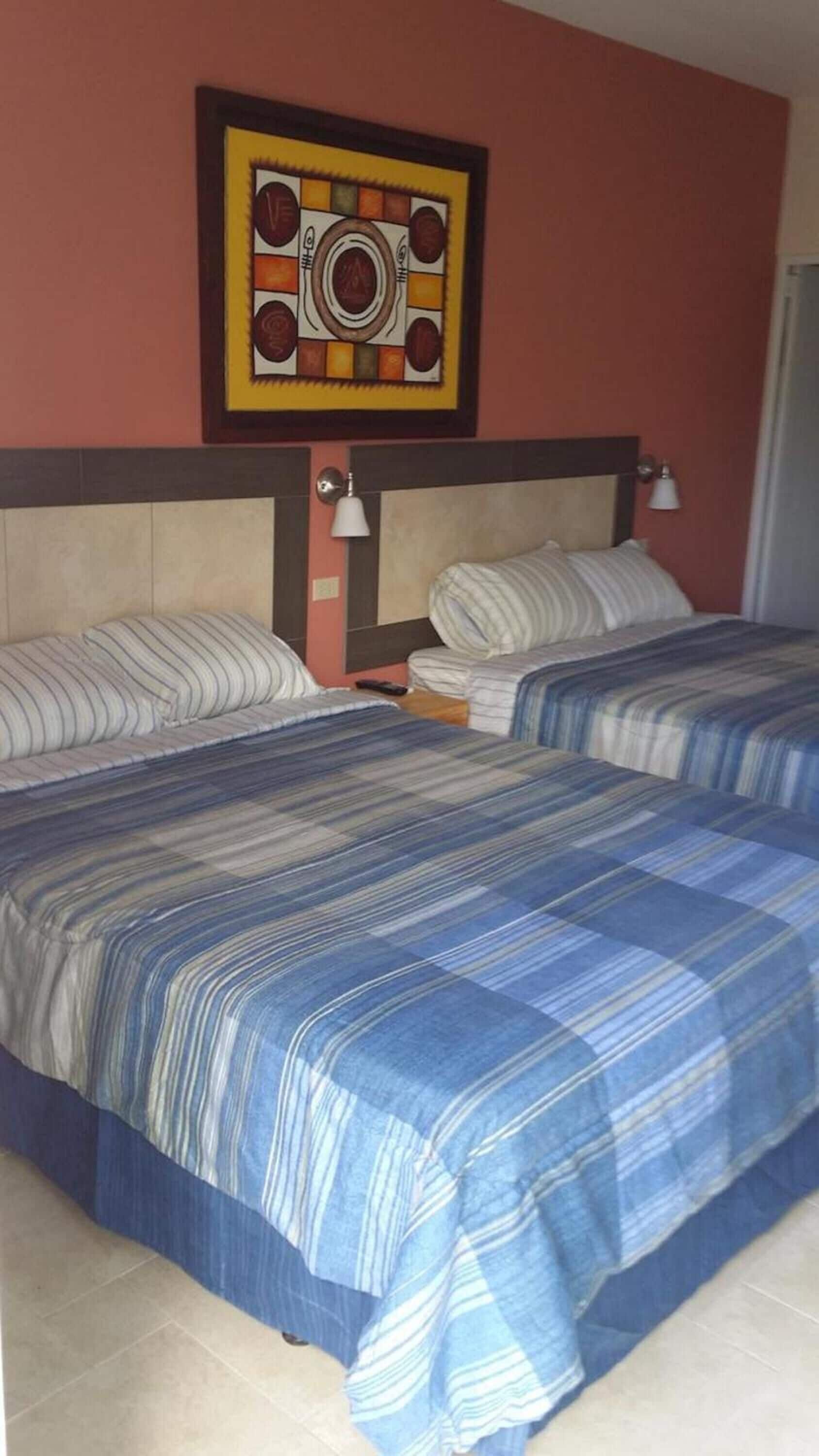 Une chambre d’hôtel avec deux lits, chacun doté de draps à rayures, d’une tête de lit avec un luminaire intégré et d’un tableau abstrait encadré au-dessus des lits.