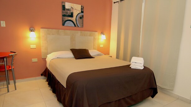 A neatly made bed with a brown bedspread and a single pillow, a small table with a red chair, and two wall-mounted lamps. - Dream's Hotel Puerto Rico (San Juan)