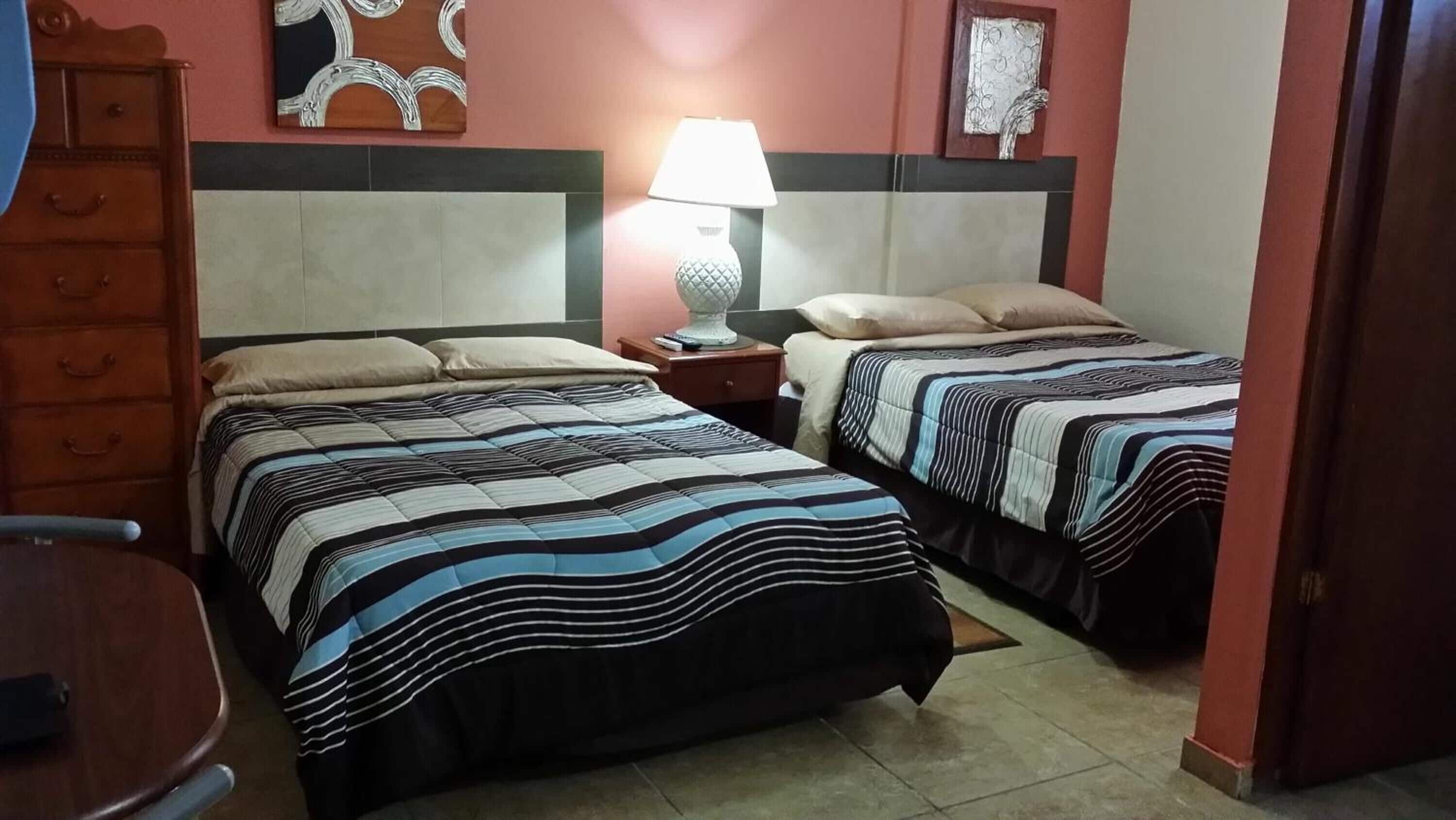 Una habitación de hotel con dos camas, una mesita de noche con lámpara, un aparador y una pared decorada con obras de arte.