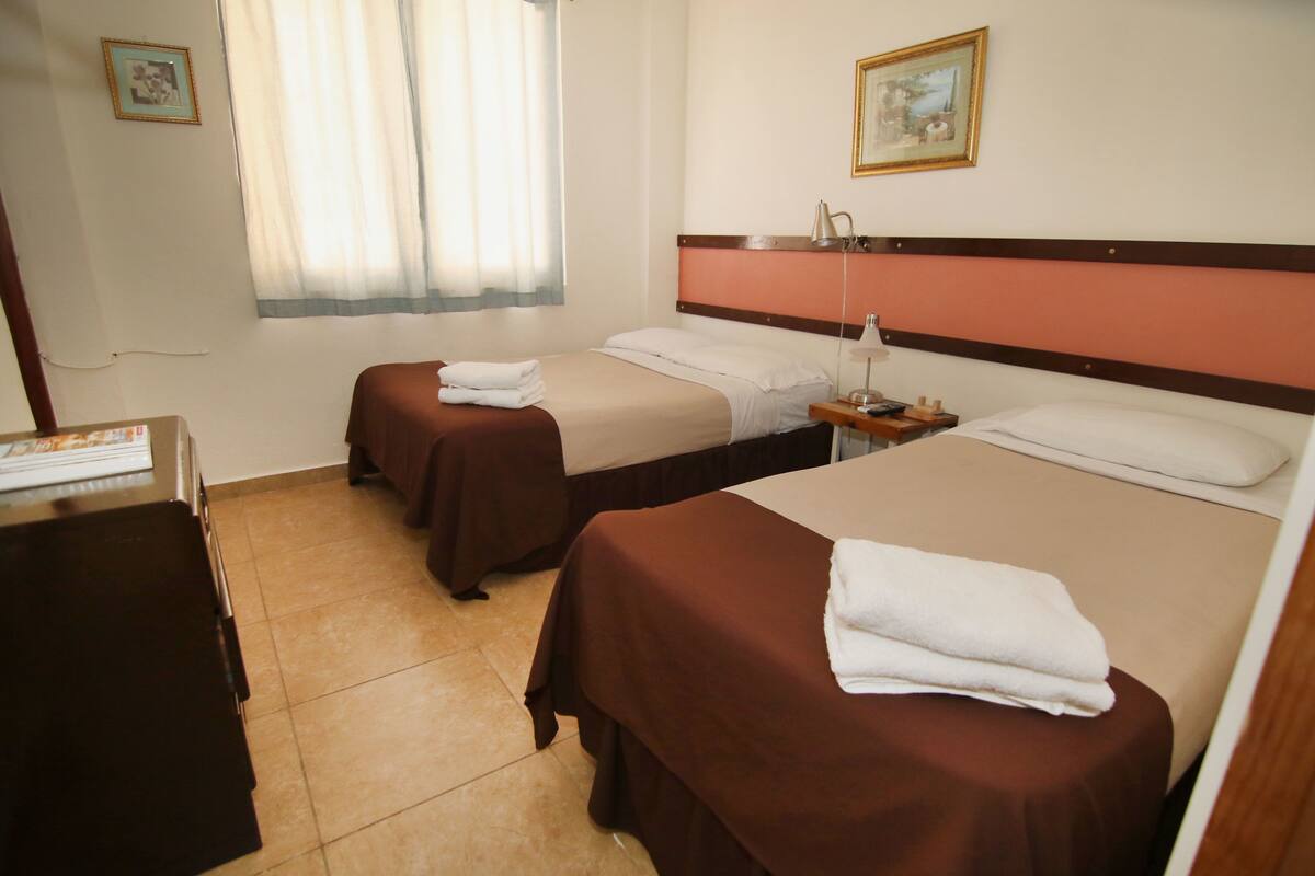 A hotel room with two beds, a desk, a lamp, and a window with curtains.