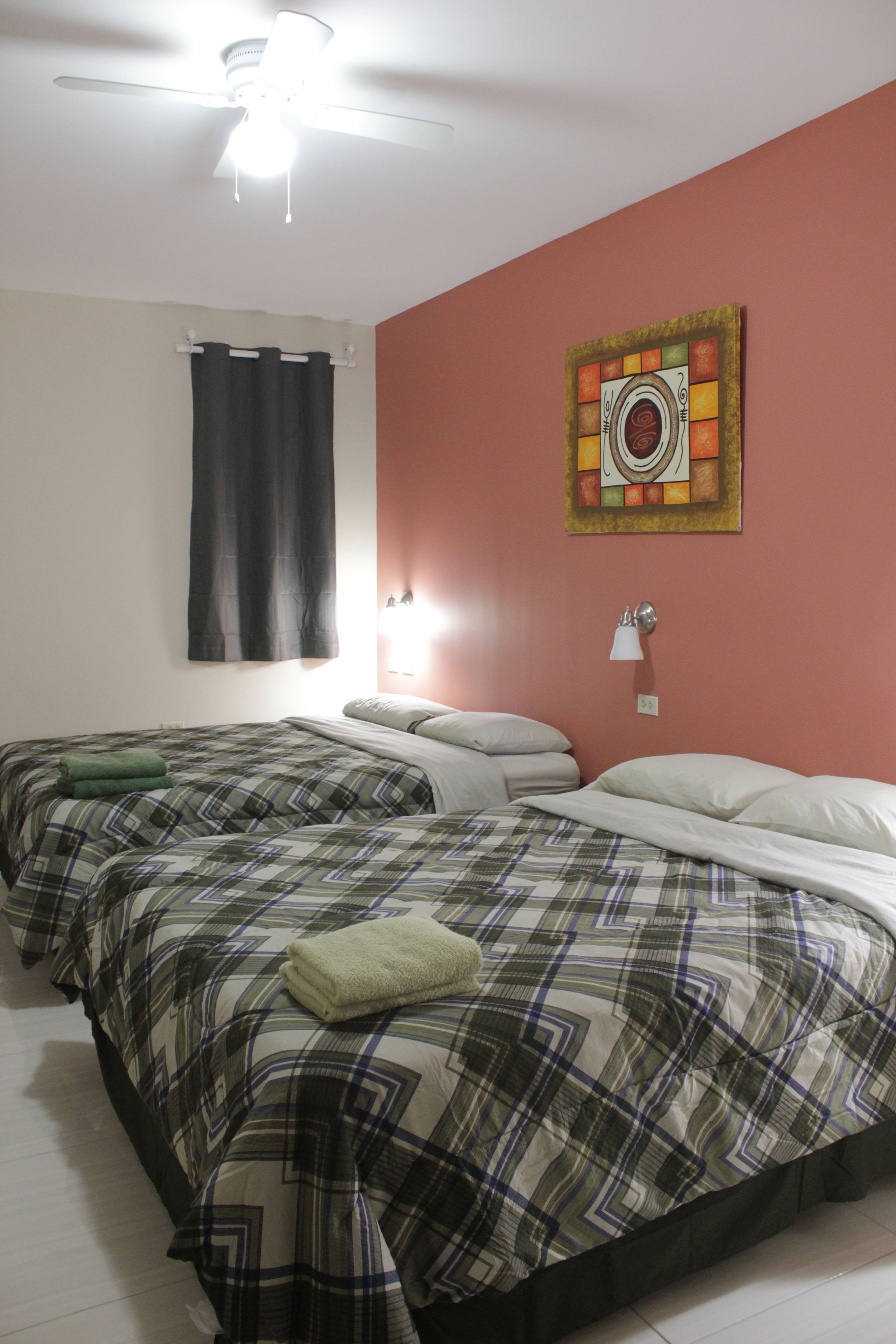 A hotel room with two beds, a ceiling fan, a painting on the wall, and a window with curtains.