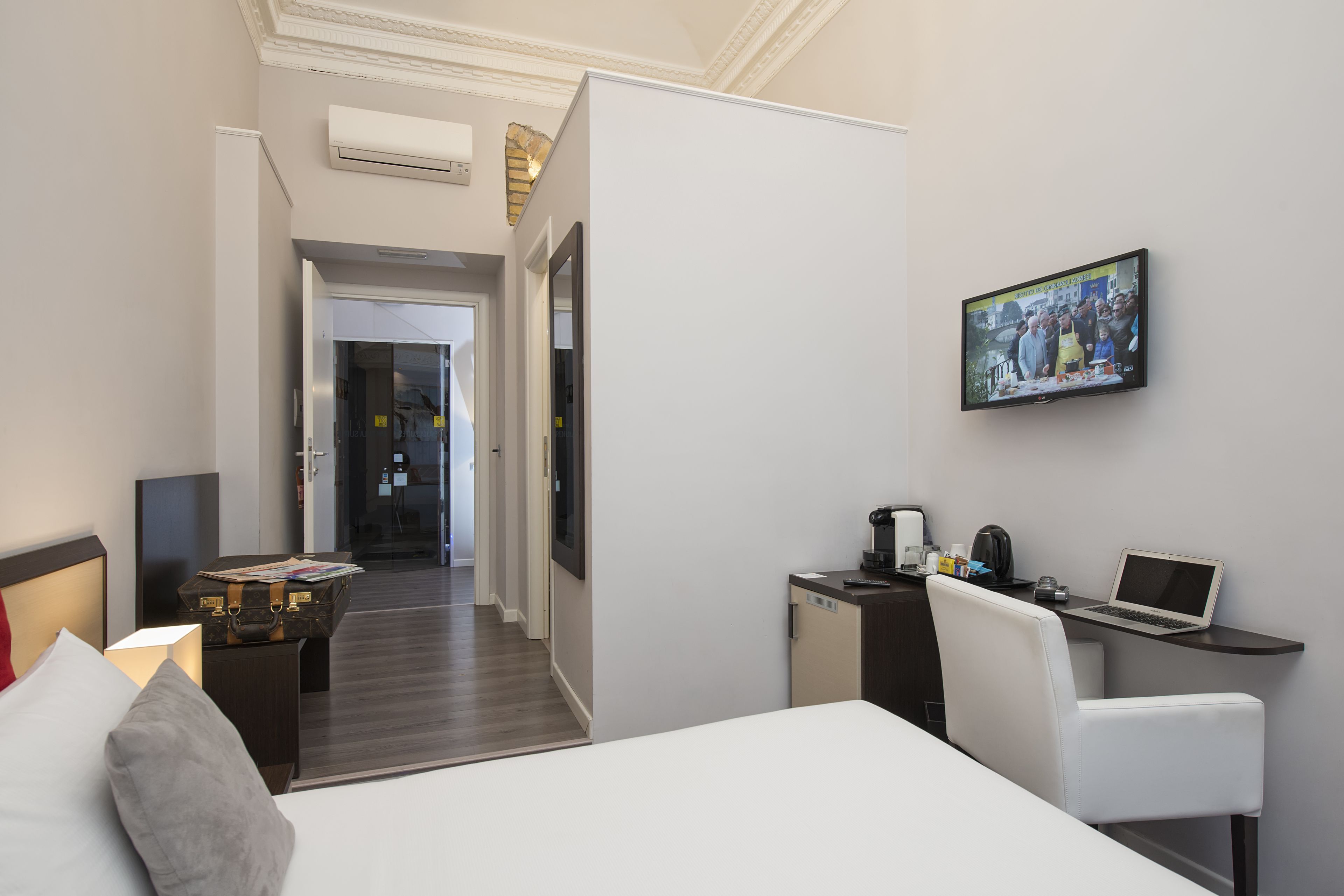 Una habitación de hotel con una cama, un escritorio con una computadora portátil, una silla, un televisor y una mesita de noche con una lámpara y una maleta.