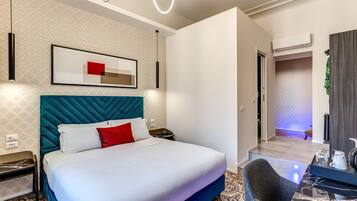 A modern hotel room with a large bed, a headboard, a bedside table, a chair, a desk with a lamp, and an air conditioning unit.