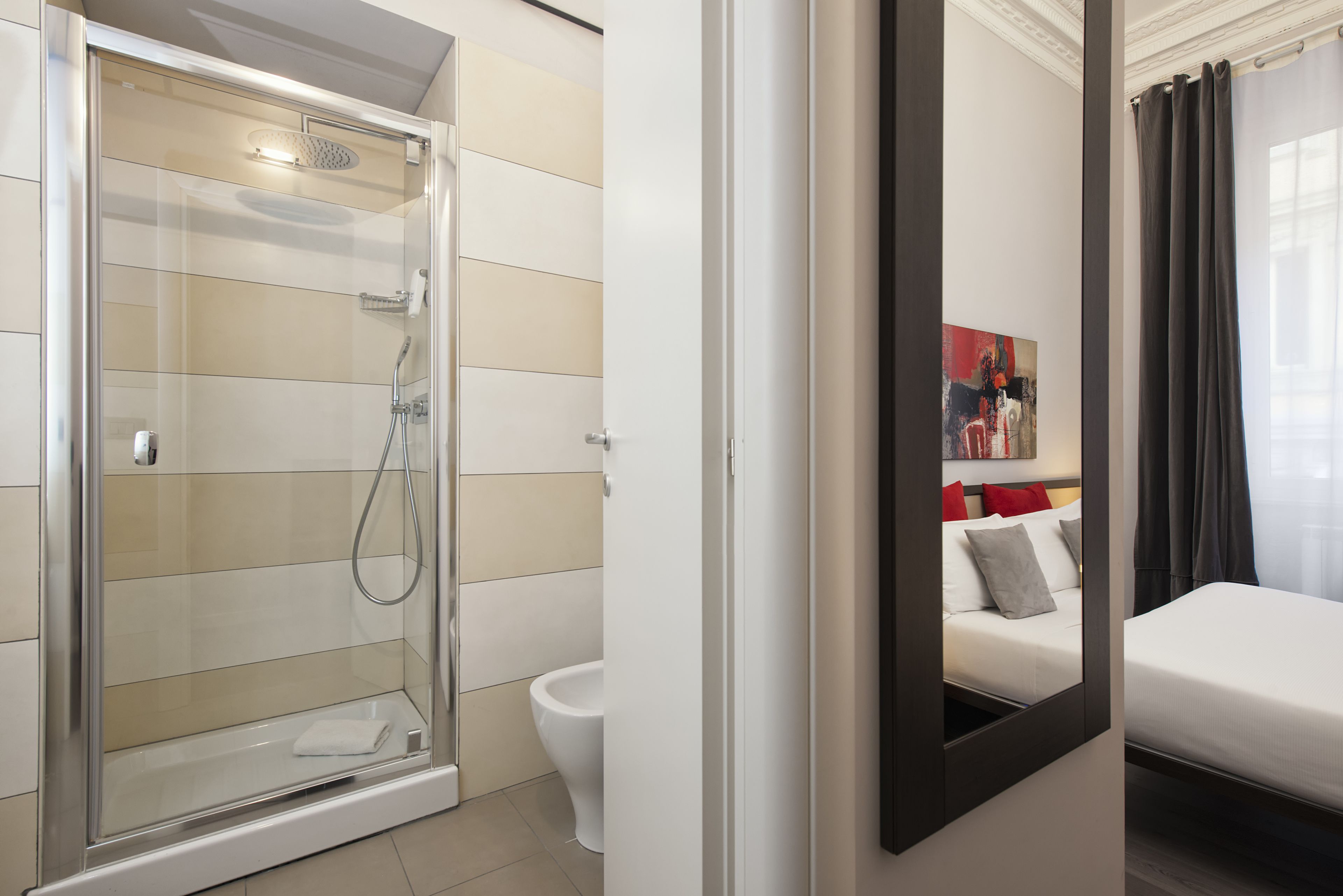 A modern bathroom with a glass-enclosed shower, a toilet, and a bed with a mirror reflecting a painting and a nightstand.