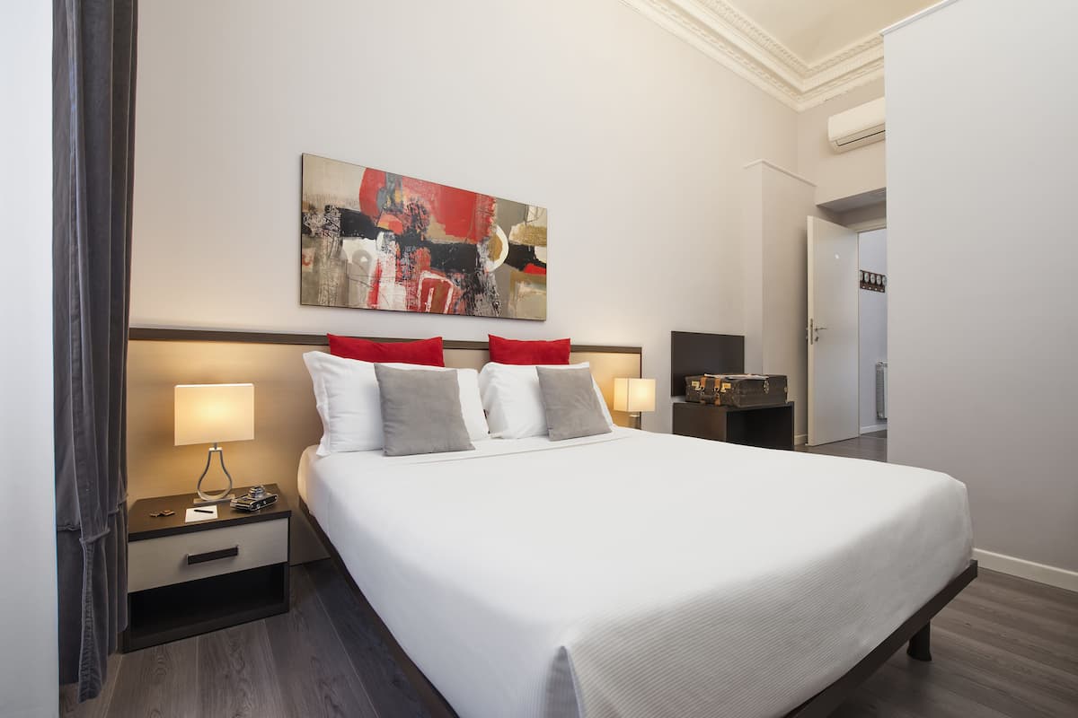 Un lit bien fait, avec des oreillers blancs et rouges, une table de chevet avec une lampe, et un tableau accroché au mur.