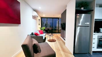 A modern apartment with a living area featuring a sofa and a dining table. A large window offers a city view.