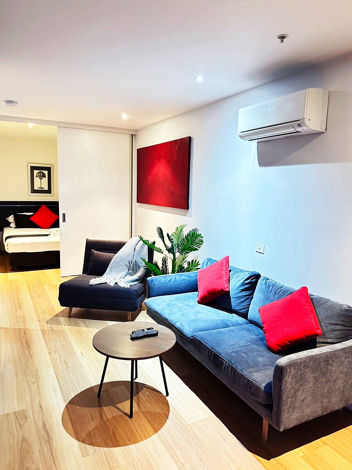 A modern living room with a sofa, armchair, and coffee table. There is a bed in the background and a large window.