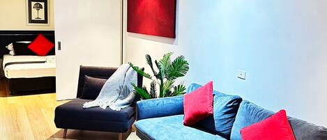 Una sala de estar moderna con sofá, sillón y mesa de centro. Se ve una cama al fondo y un ventanal grande.