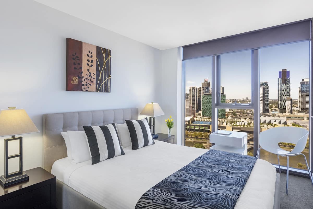 Una habitación de hotel moderna con una cama grande, vistas a la ciudad, una silla blanca y una mesita de noche con lámpara.