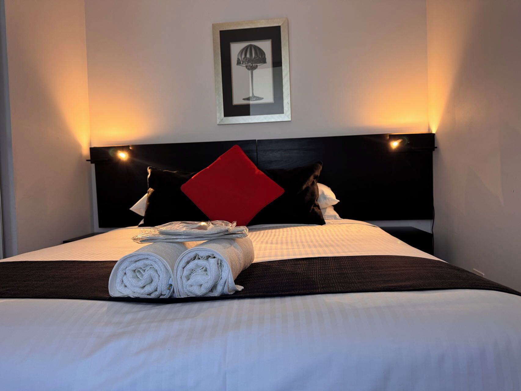 Una cama bien hecha con toallas enrolladas, una almohada roja y un cuadro en la cabecera.