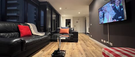 Una sala de estar moderna con un sofá de cuero negro, una mesa de centro de vidrio y un televisor montado en la pared.