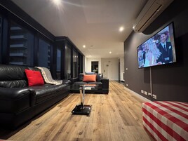 A modern living room with a black leather sofa, a glass coffee table, and a TV mounted on the wall.