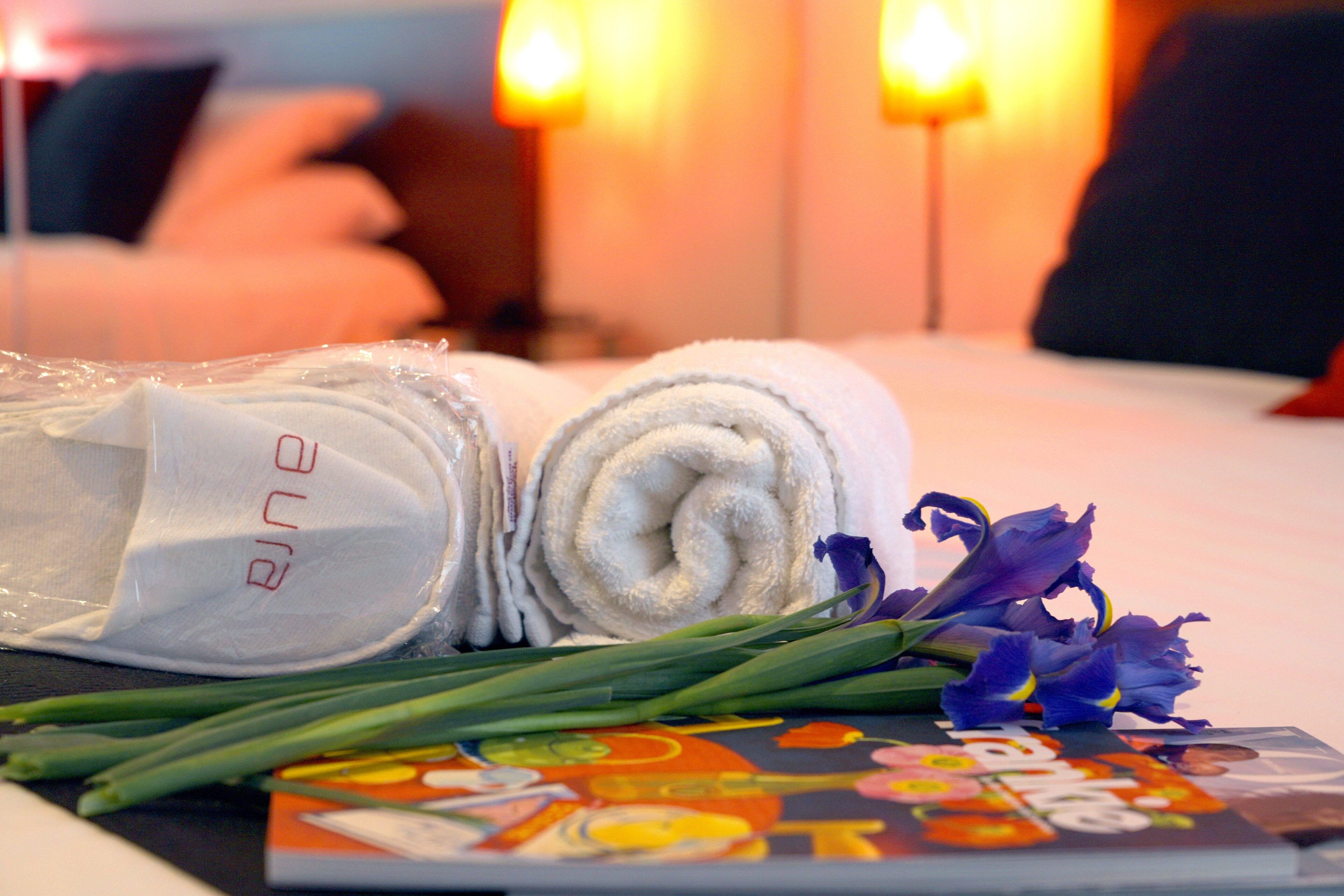 A neatly made bed with rolled white towels, a bouquet of purple flowers, and a magazine on a table.