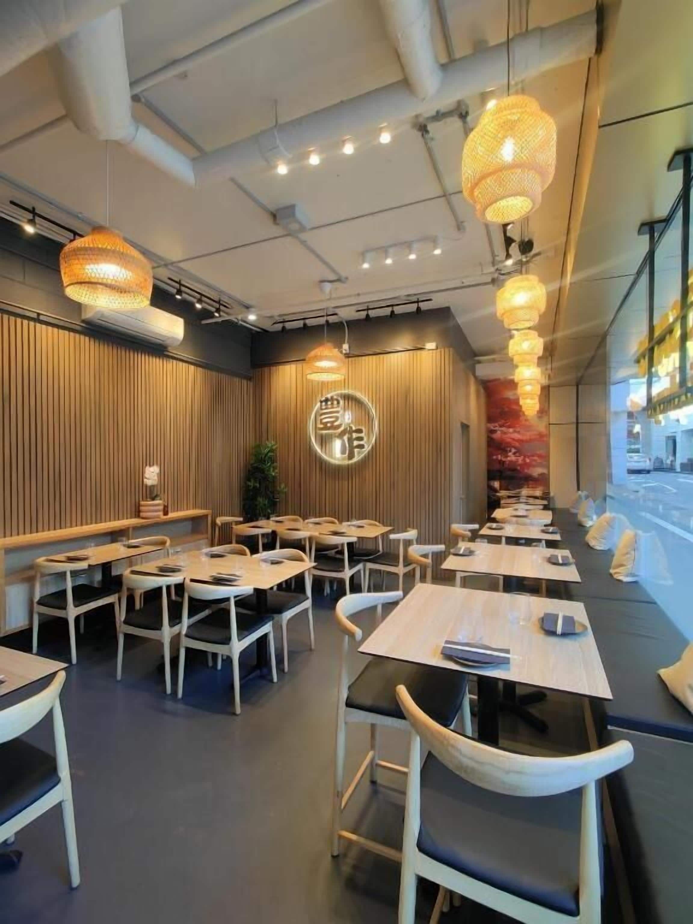 A modern restaurant interior with wooden panel walls, multiple dining tables, and hanging pendant lights.