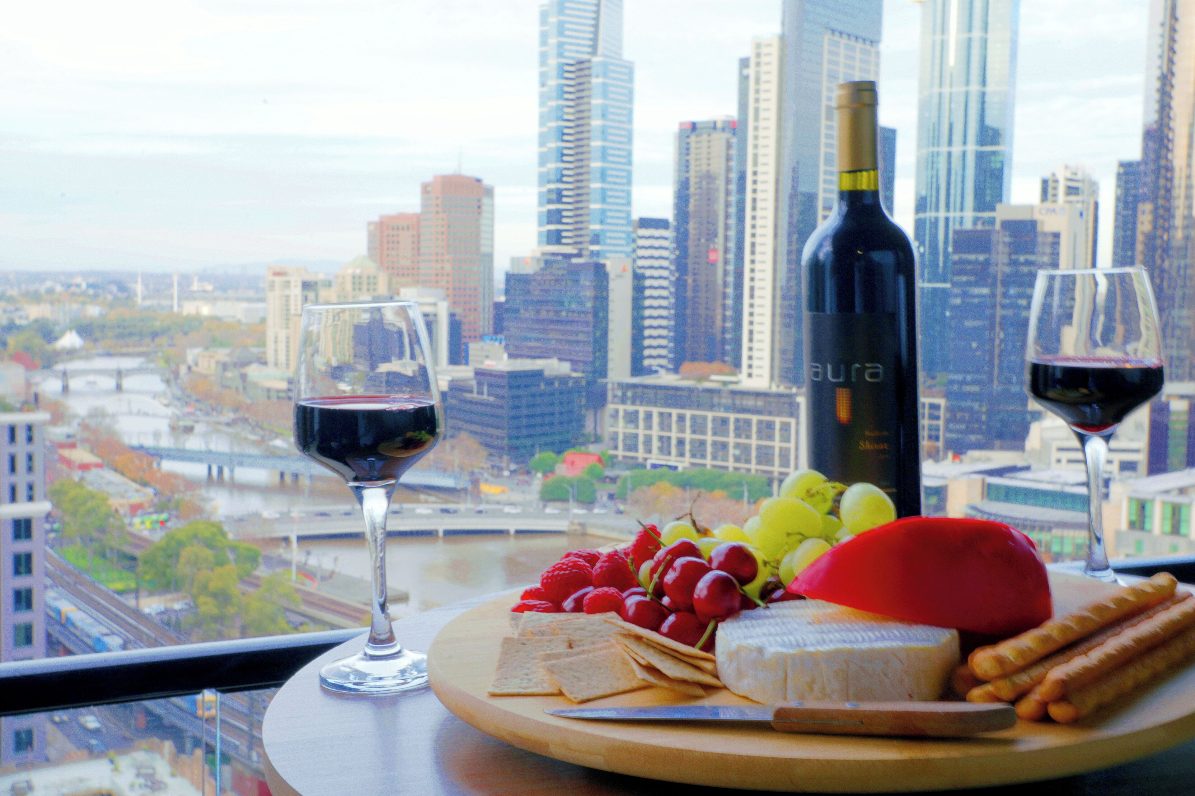 A cityscape view from a high-rise window with a table featuring a bottle of wine, two glasses, cheese, grapes, strawberries, and crackers.