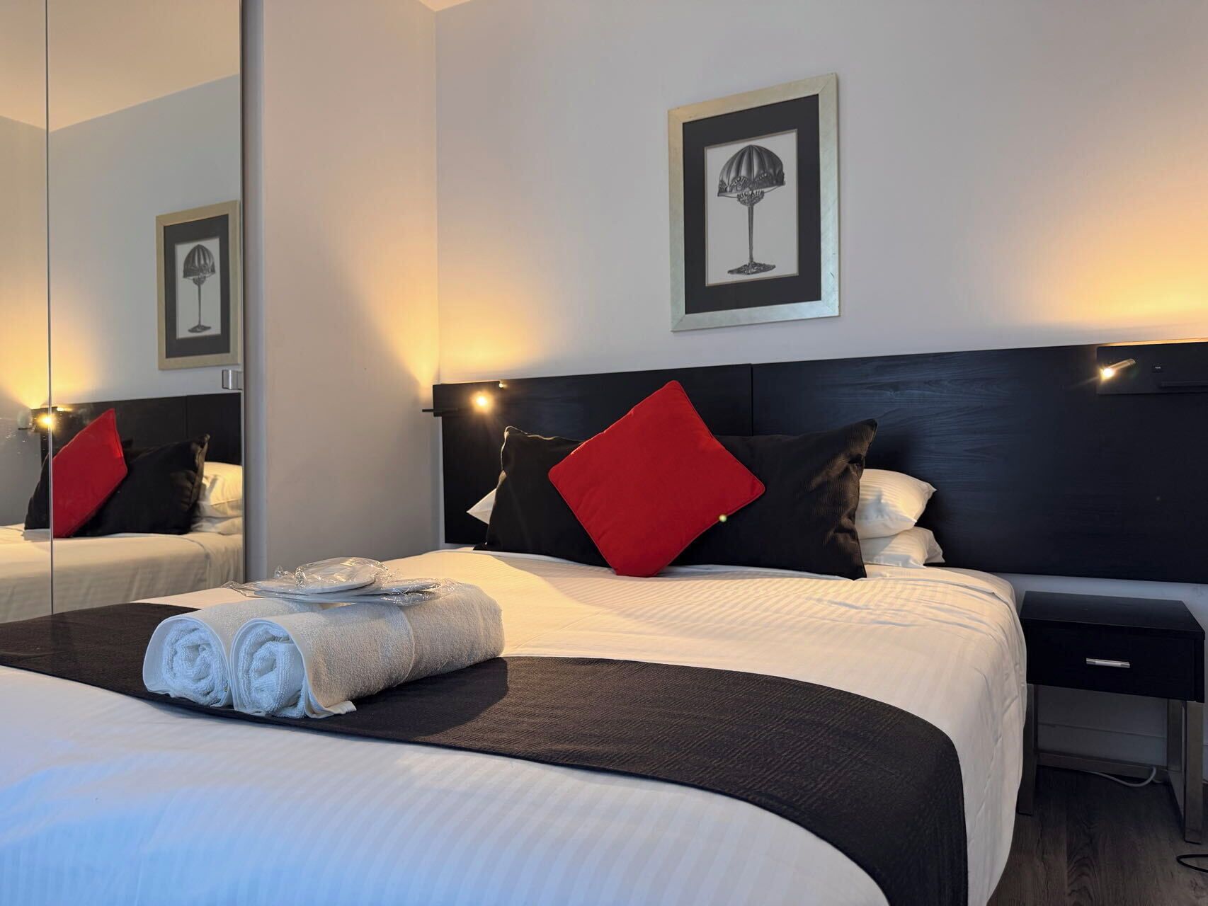 A hotel room with a large bed, a bedside table, a desk, and a mirror.