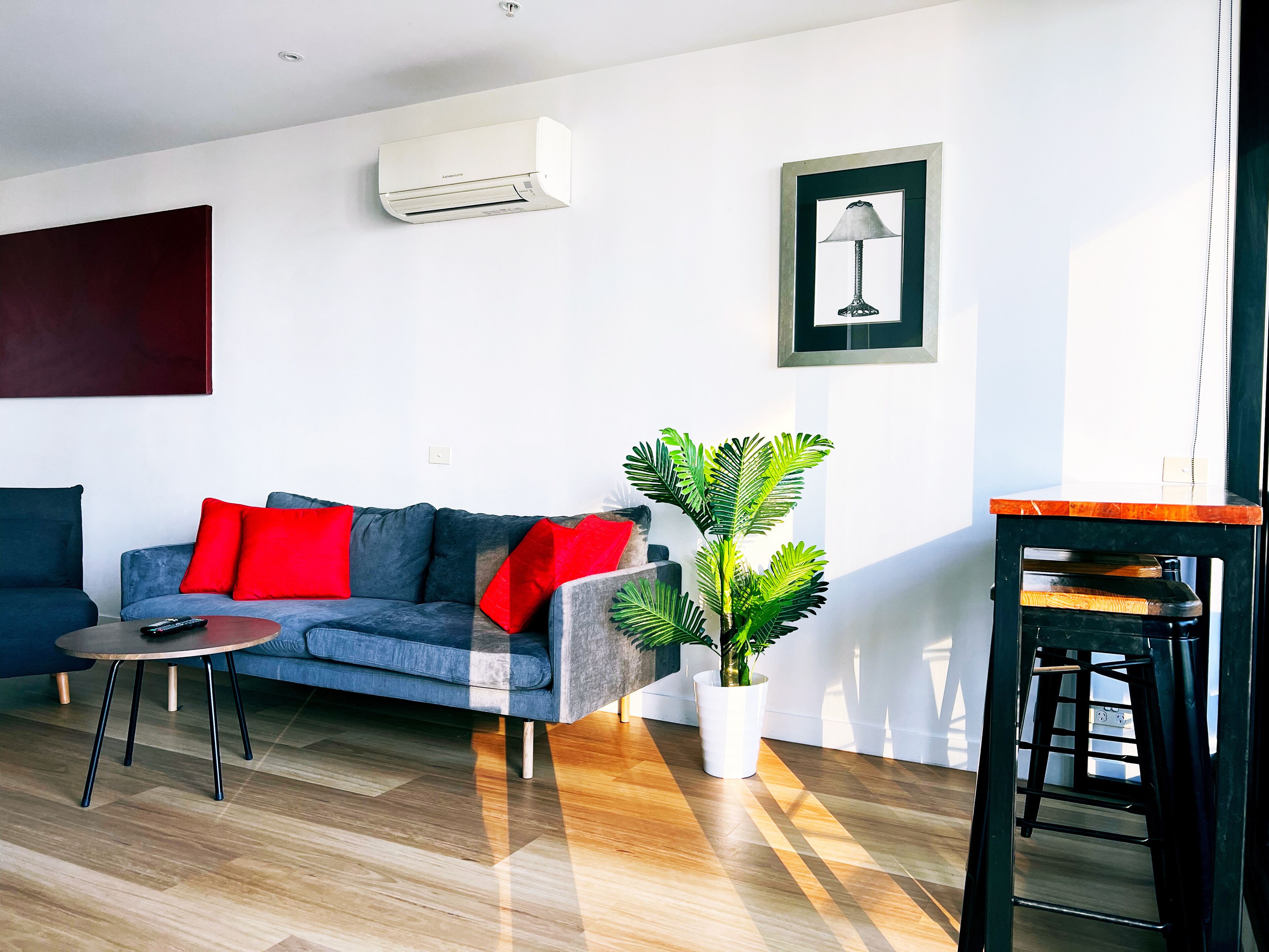 A modern living room with a blue sofa, red cushions, a small table, a potted plant, and a black stool.
