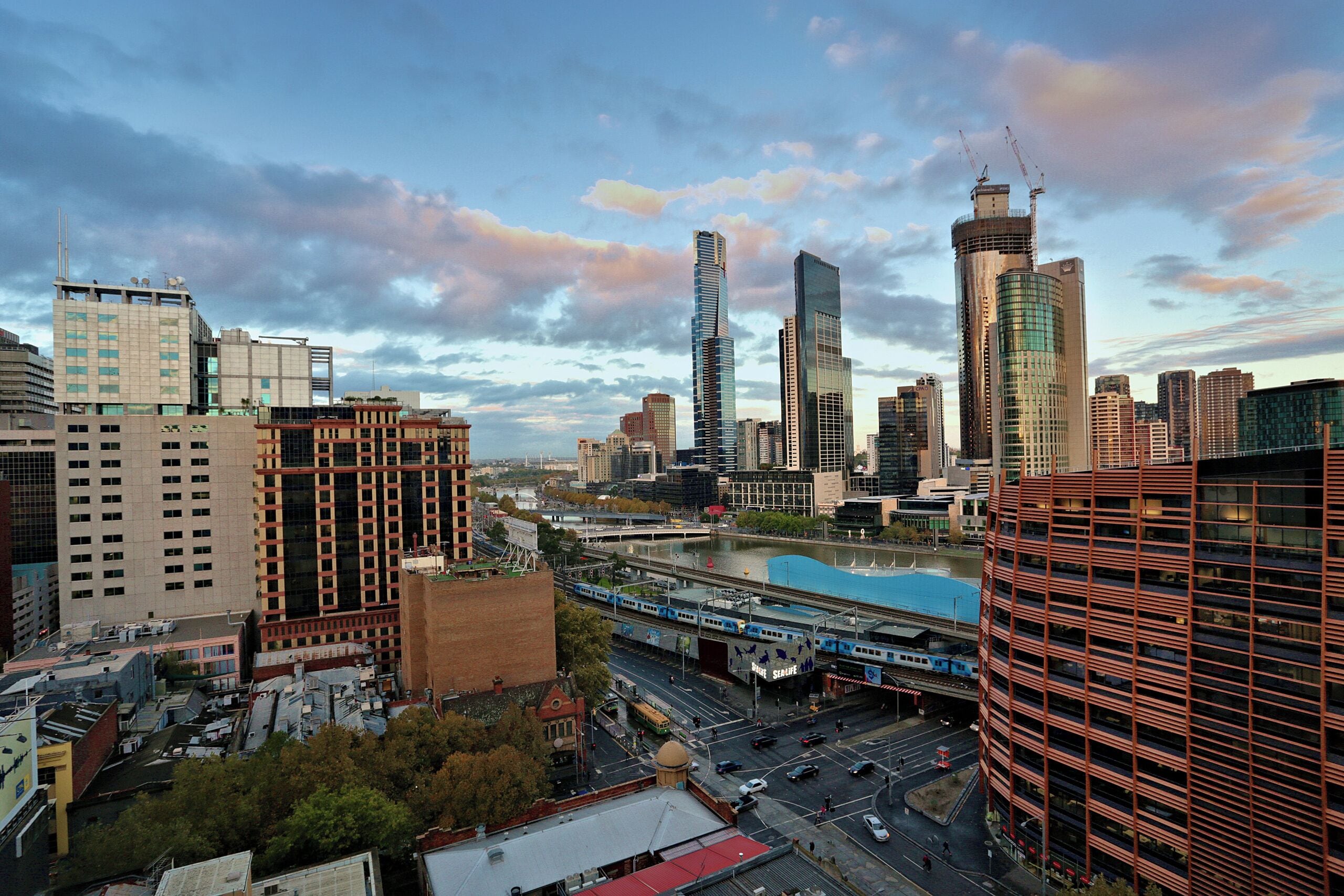 A cityscape with modern high-rise buildings, a river, and a train passing underneath a bridge.