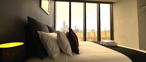 A hotel room with a large bed, a nightstand, a lamp, and a city view through large windows.