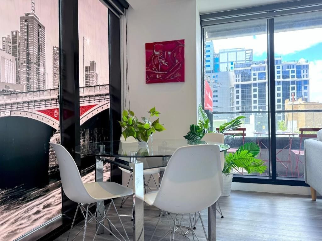 A modern living room with a glass table, white chairs, and a view of a cityscape through large windows.