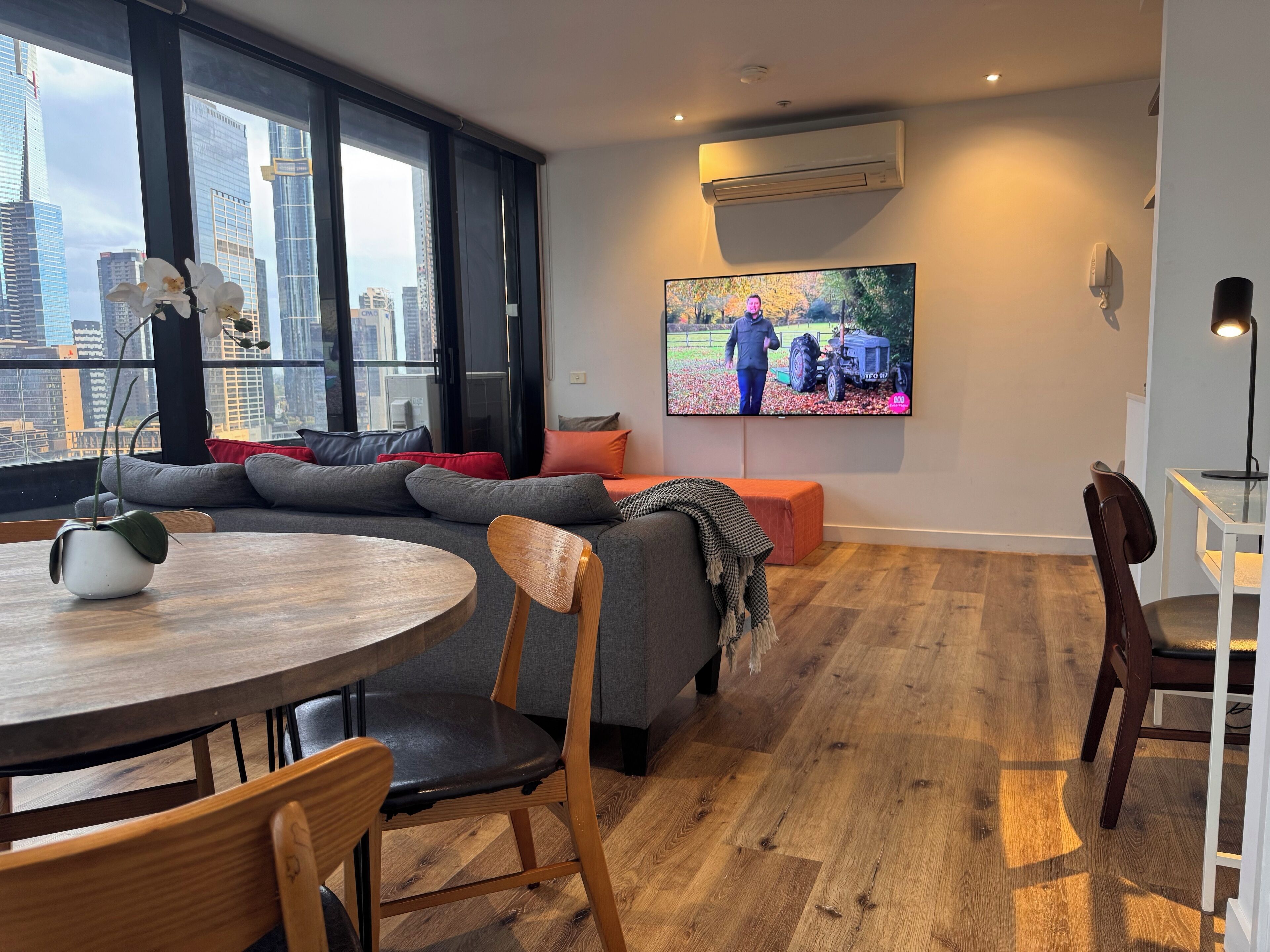Una sala de estar moderna con un ventanal grande, un televisor de pantalla plana, un sofá gris y una mesa de comedor de madera.