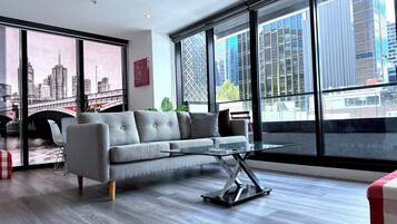 A modern living room with a grey sofa, a glass coffee table, and large windows offering a city view.