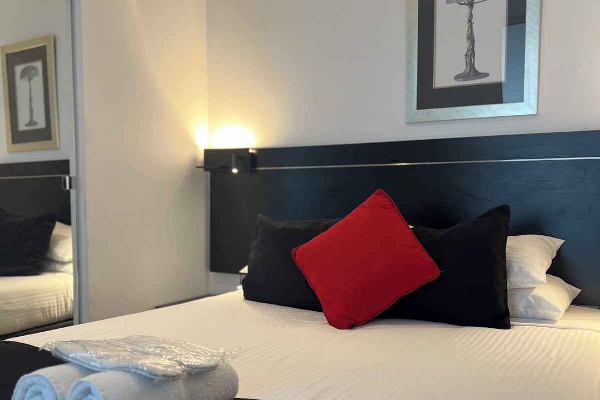 Una cama bien hecha con ropa de cama blanca y negra, una almohada roja y toallas enrolladas sobre un cabecero blanco.