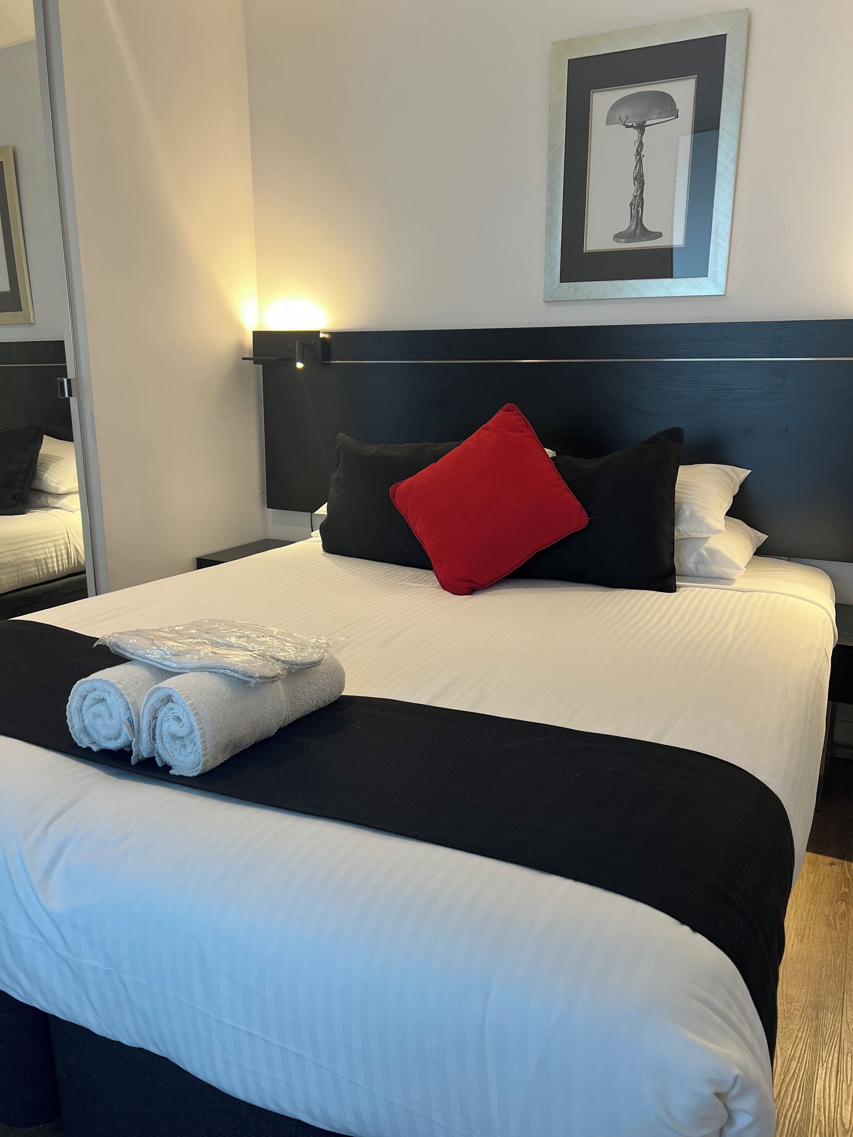 Una cama bien hecha con ropa de cama blanca y negra, un cojín rojo y toallas enrolladas sobre una bandeja negra.