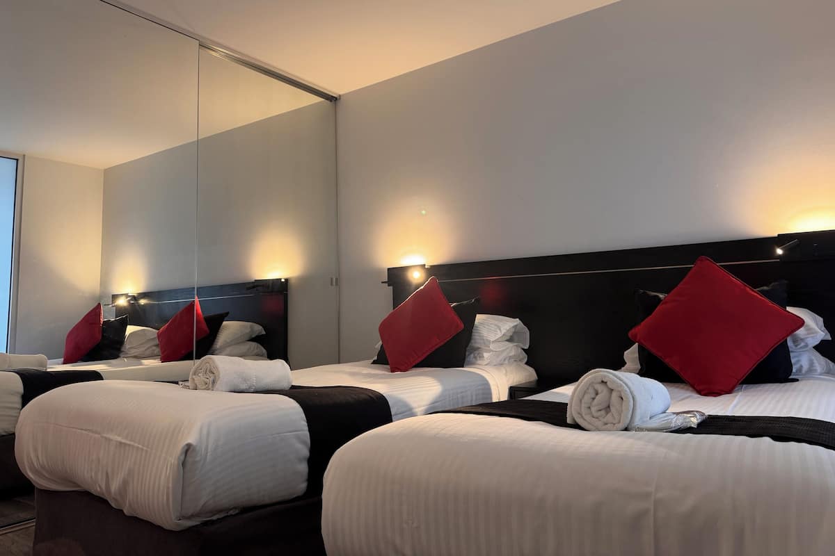 Habitación de hotel con cuatro camas dispuestas en fila, cada una con ropa de cama blanca y almohadas rojas.