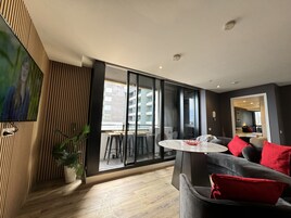 A modern living room with a large sliding glass door leading to a balcony, a round table, and a grey sofa with red cushions.