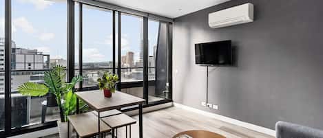 A modern living room with a large window, a flat-screen TV, a wooden coffee table, a grey sofa, and a potted plant.