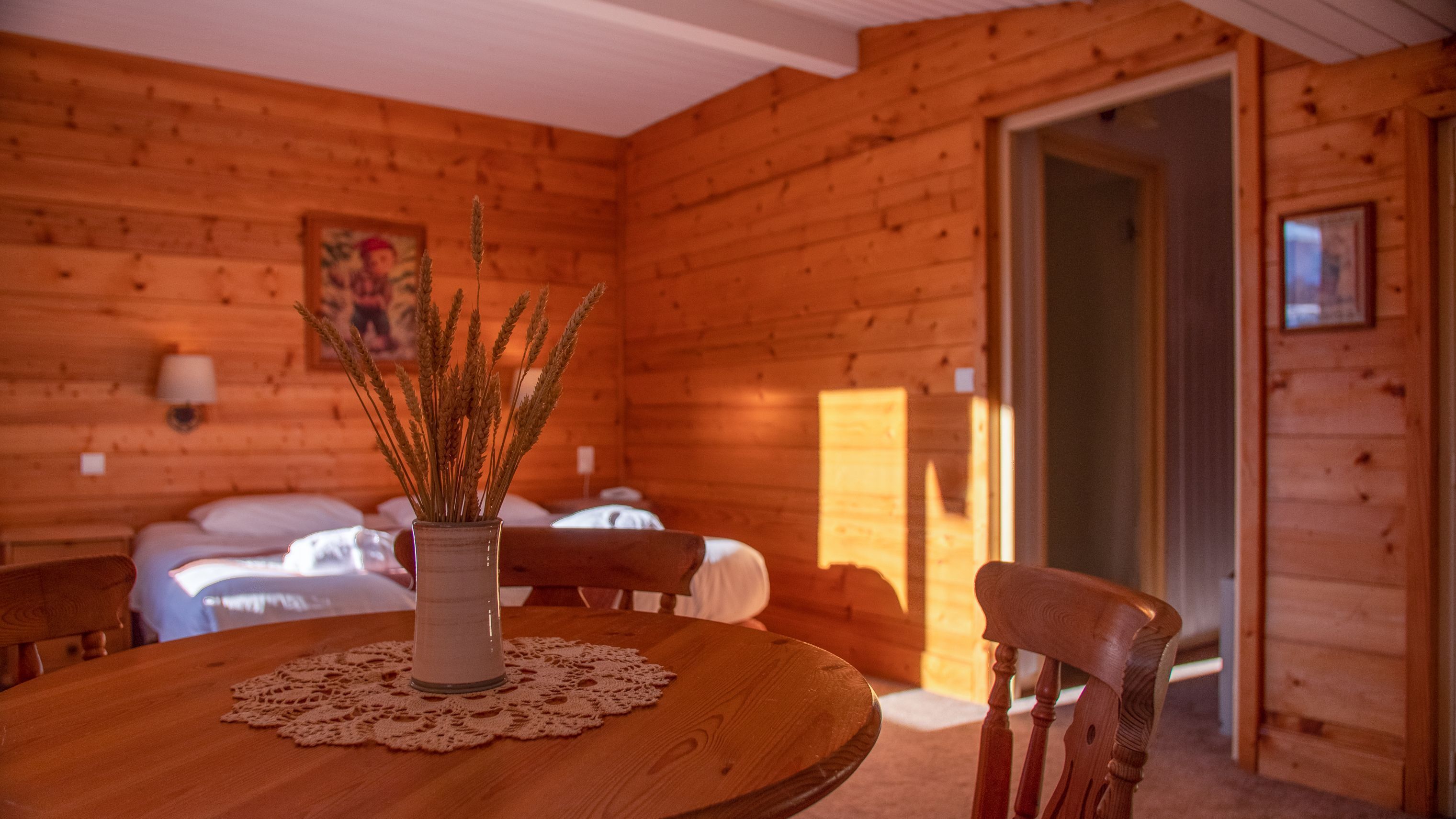 Interior de una cabaña de madera con una mesa de comedor, sillas, un florero con espigas de trigo y una cama con ropa blanca.