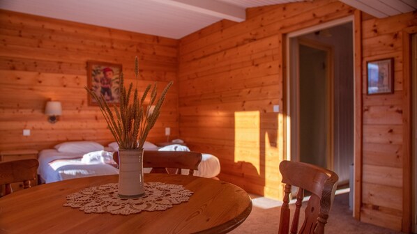 A wooden cabin interior with a dining table, chairs, a vase with wheat stalks, and a bed with white linens.