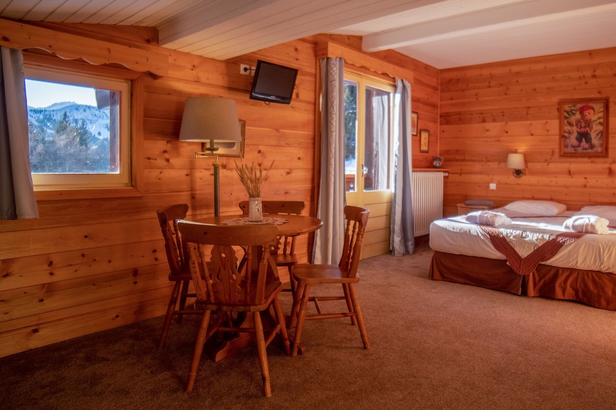 A cozy wooden cabin room with a bed, a dining table with chairs, a TV mounted on the wall, and a window with curtains.