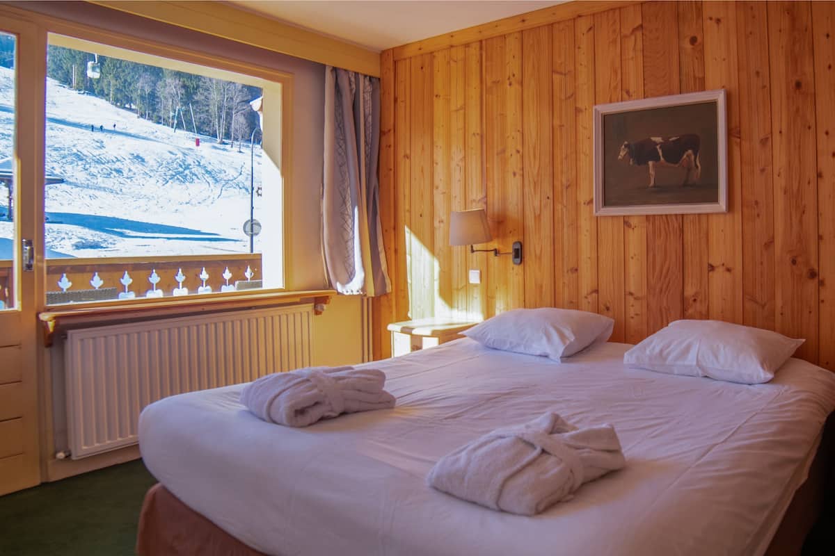 A hotel room with a wooden paneled wall, a bed with white linens, two bedside lamps, a framed picture of a cow, and a window with a view of a snowy landscape.