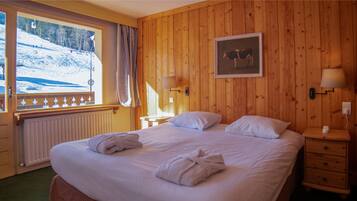 A hotel room with a wooden paneled wall, a bed with white linens, two bedside lamps, a framed picture of a cow, and a window with a view of a snowy landscape.