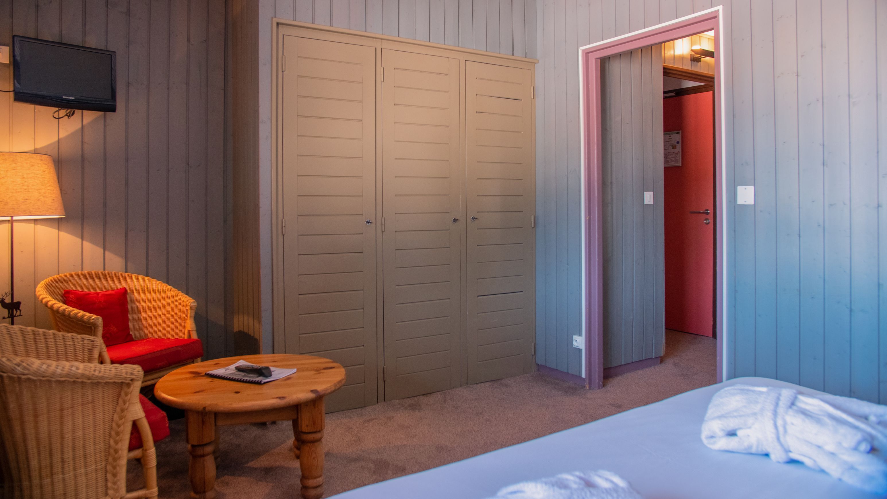 A room with a bed, a wooden table, a wicker chair, a wardrobe, and a television mounted on the wall.