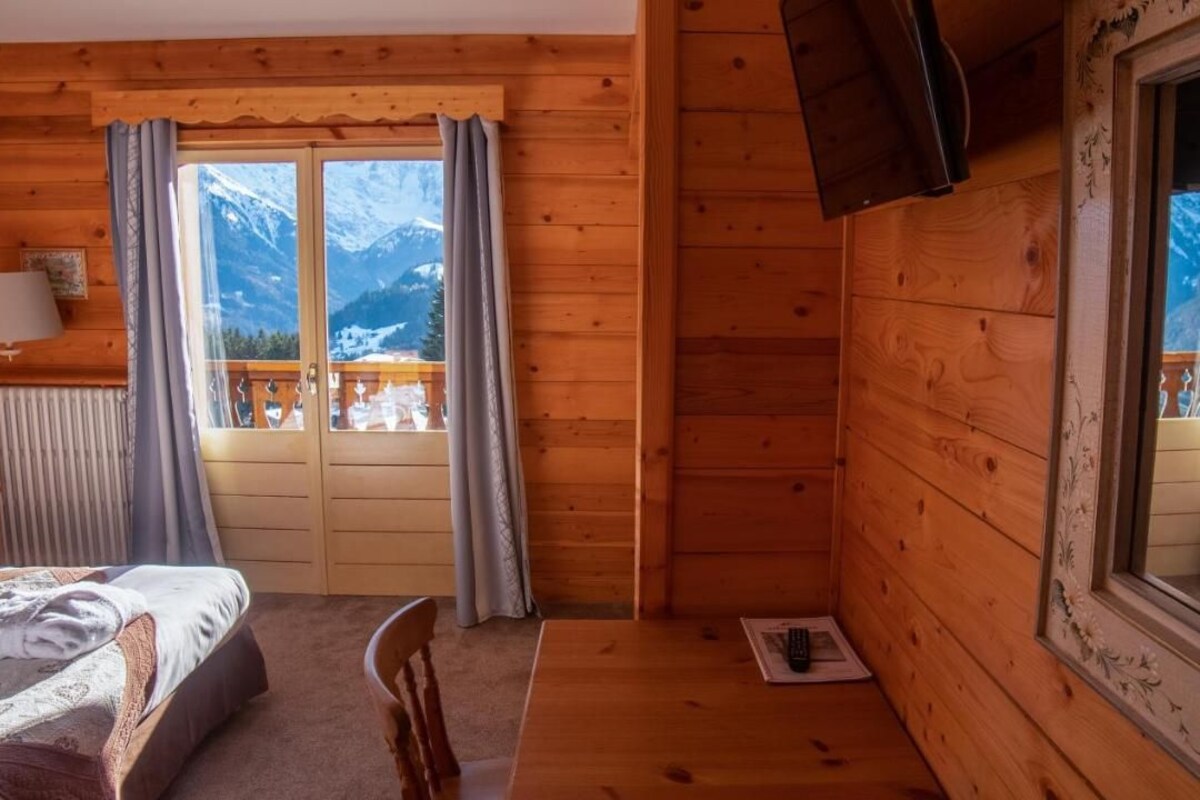 A wooden cabin room with a bed, a table, a chair, a window with curtains, and a view of snow-capped mountains.
