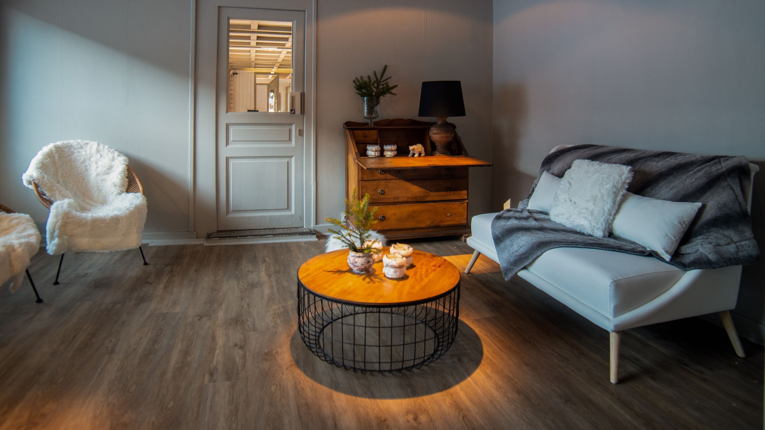 Una sala de estar con un sillón blanco, un sofá con una manta de piel, una mesa de centro de madera con una planta y un aparador de madera con una lámpara.