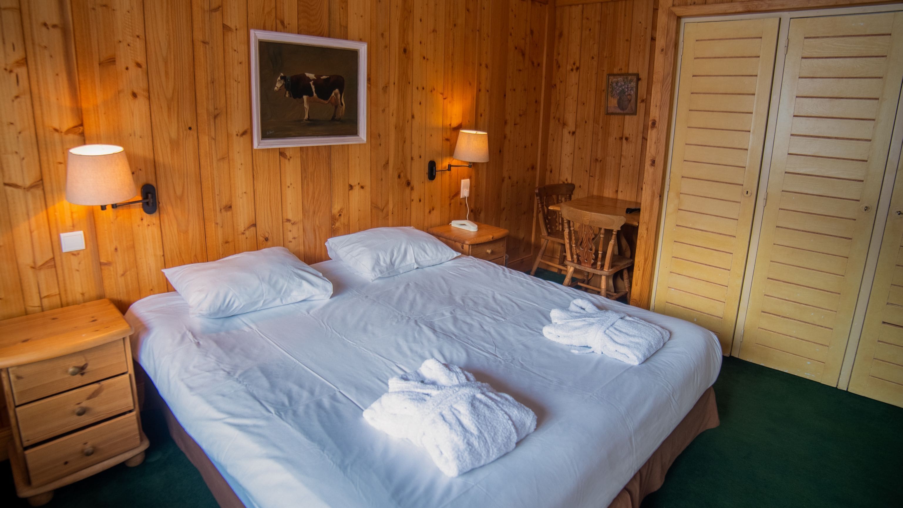 A wooden-paneled room with a large bed, two bedside lamps, a framed picture, a small table, and a chair.
