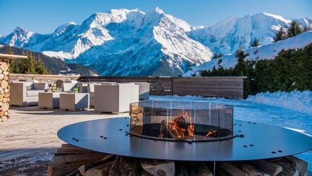 A circular outdoor seating area with a fire pit in the center, surrounded by stone walls and a view of snow-capped mountains. - Hôtel Arbois Bettex (Saint-Gervais-les-Bains)