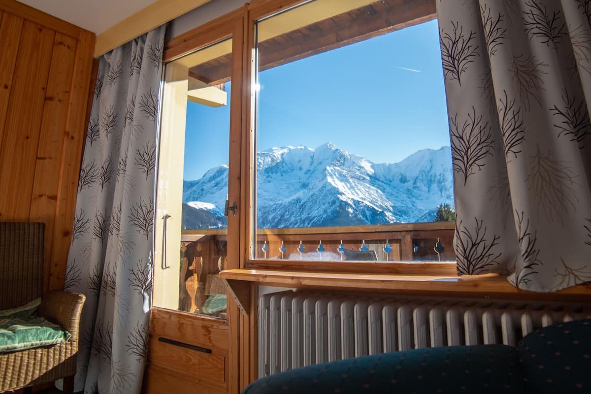A room with a wooden interior, a window with a view of snow-capped mountains, a wicker chair, and a radiator.