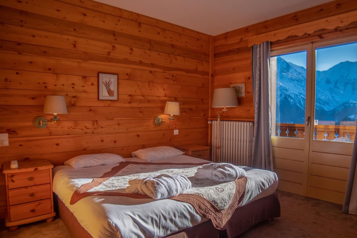 A wooden cabin room with a bed, two bedside lamps, a framed picture, and a view of mountains through a large window.