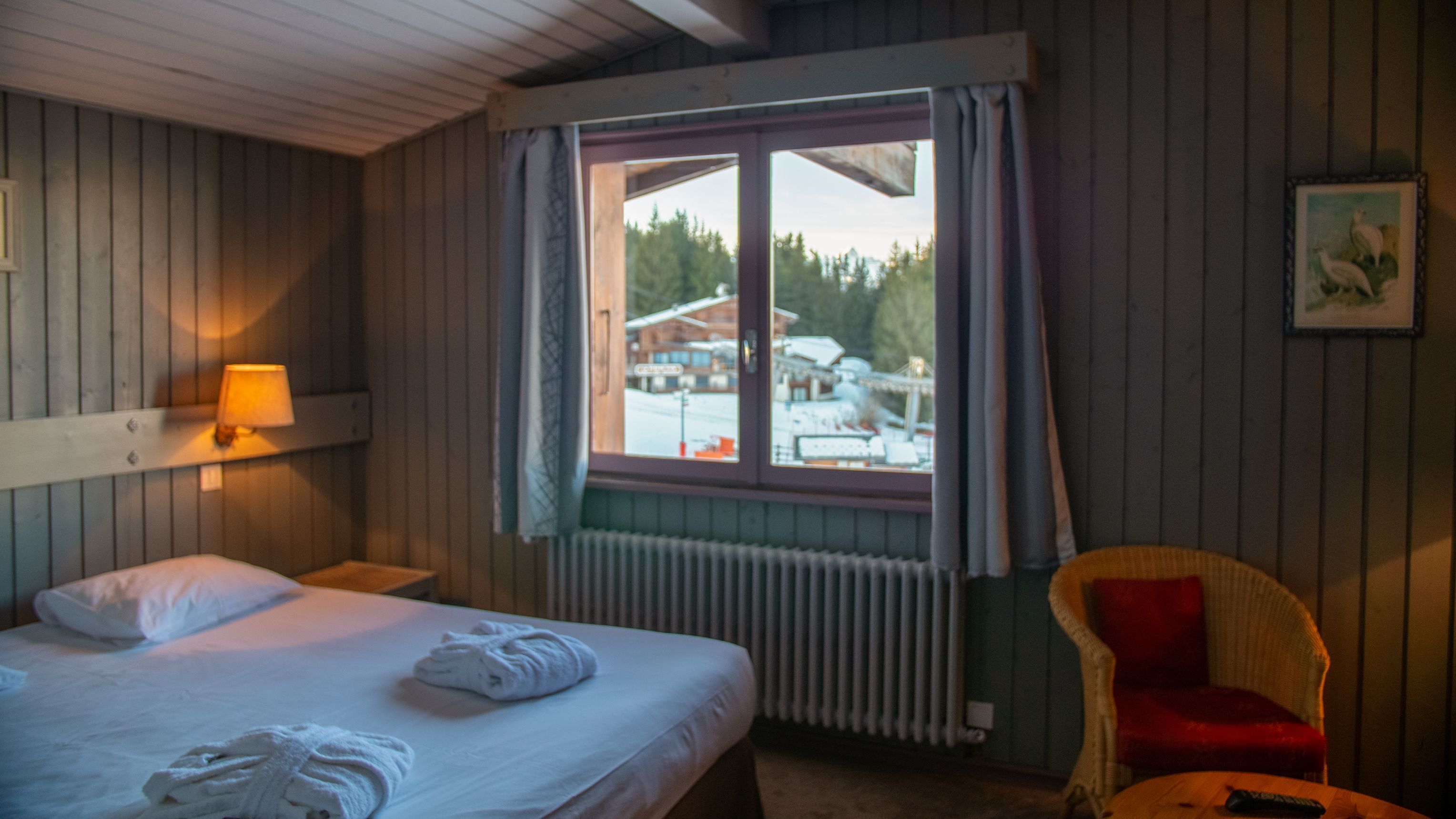A hotel room with a bed, a chair, a window with a view of a building and trees, and a lamp on the wall.