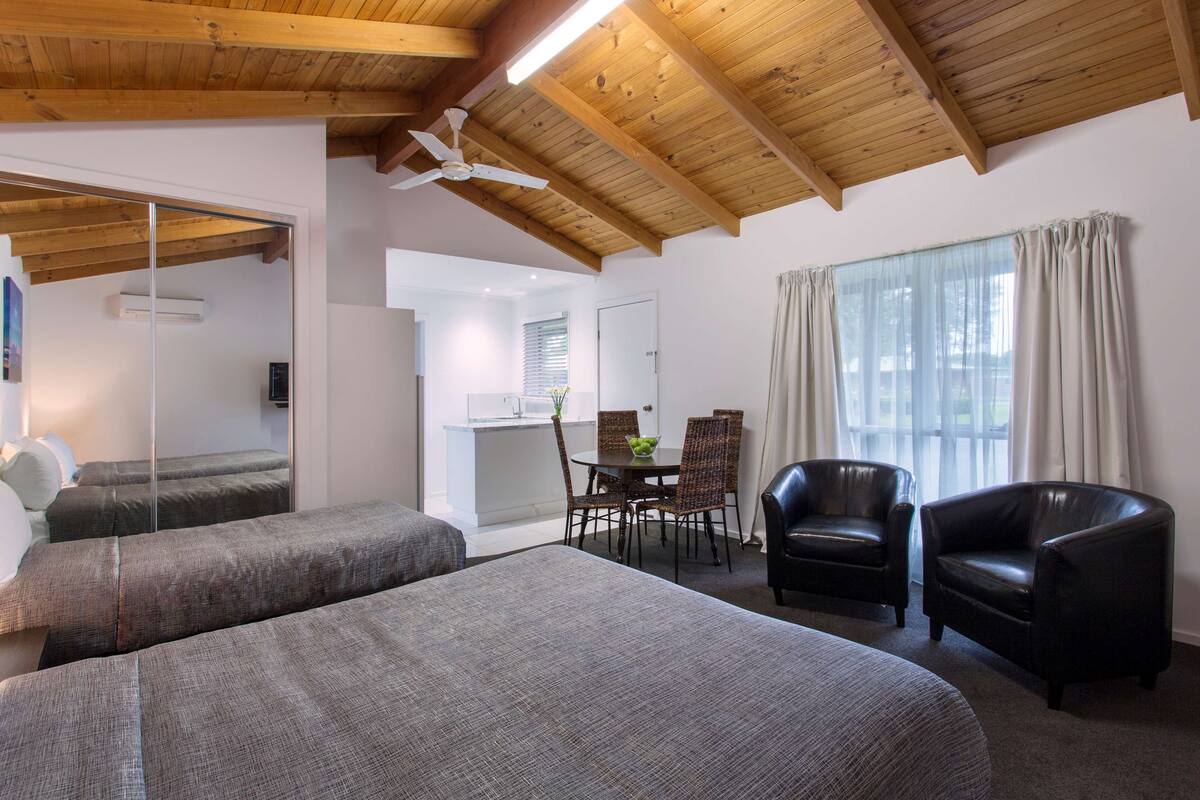 A room with two beds, a dining table with chairs, a ceiling fan, and large windows with curtains.