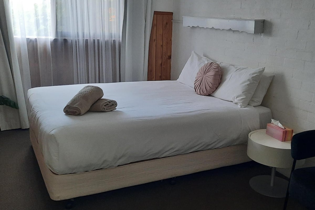 A neatly made bed with rolled towels, a bedside table with a tissue box, and a chair.