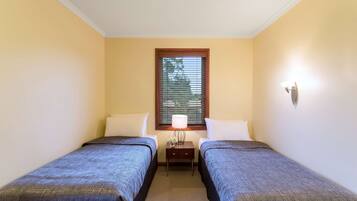A small, well-lit room with two single beds, a nightstand with a lamp, and a window with blinds.