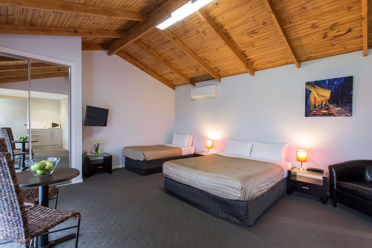 A hotel room with two beds, a dining area with a table and chairs, a TV mounted on the wall, and a painting above the beds.